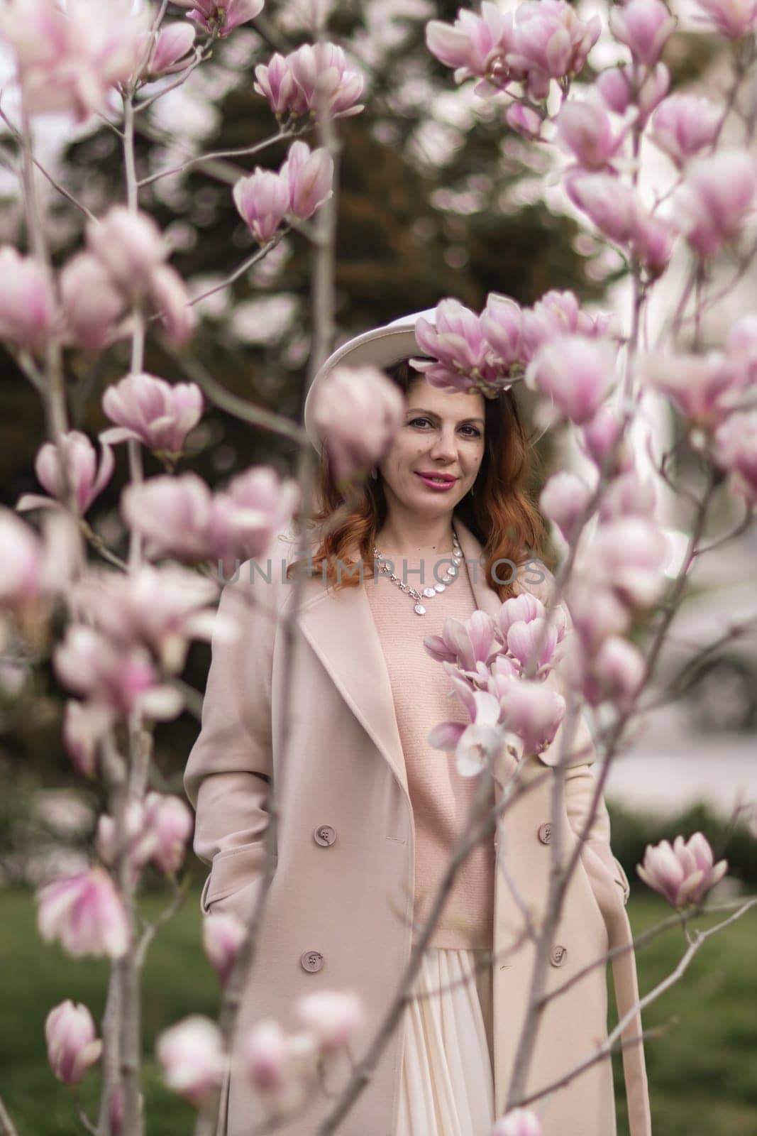 Woman magnolia flowers, surrounded by blossoming trees., hair down, white hat, wearing a light coat. Captured during spring, showcasing natural beauty and seasonal change. by Matiunina