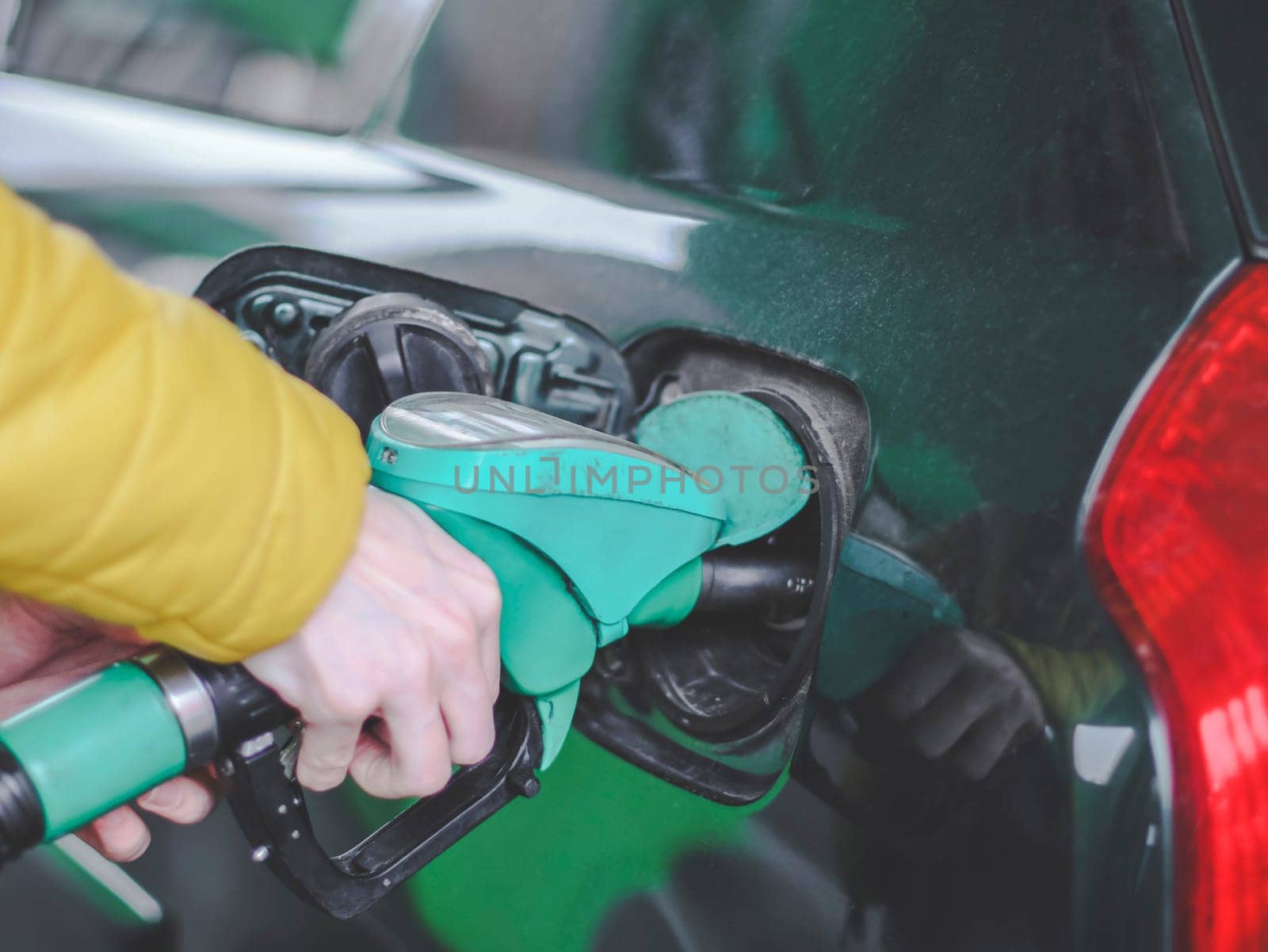 The hand of a young caucasian man in a yellow jacket holds a gas station gun and refuels a car with gasoline at a city gas station, close-up side view. Gas station concept, service.