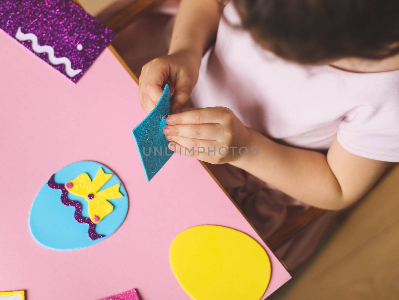 Hands of a little caucasian girl holding a blue sticker and peeling off a hat for felt eggs, sitting at a children's table with a set of handicrafts on a pink background, flat lay close-up.Craft concept,diy,needlework,at home,artisanal,children art,children creative,preparation Easter.