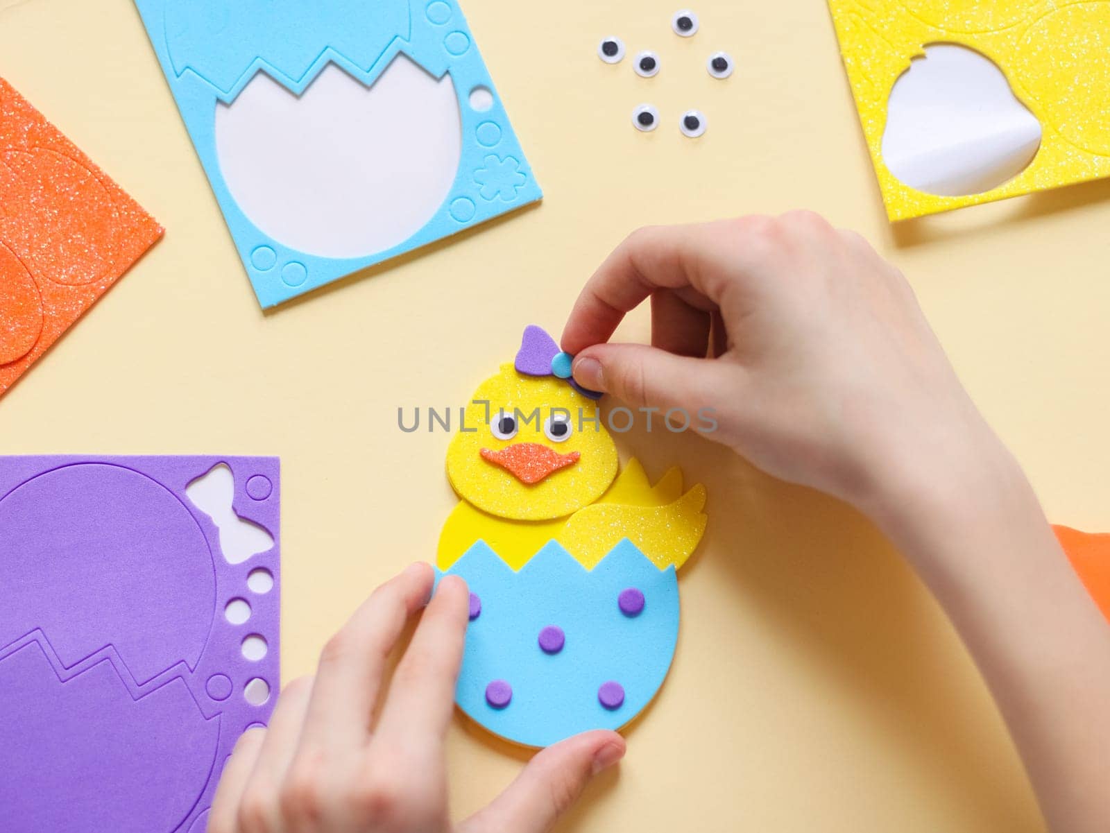 The hands of a caucasian teenage girl stick a lilac bow sticker on a yellow chicken felt with her fingers, sitting at a children's table with a set of handmade items on a pale yellow background with selective focus, flat lay closeup. The concept of crafts, diy, needlework, diy, children art, artisanal, Easter preparation,children creative.