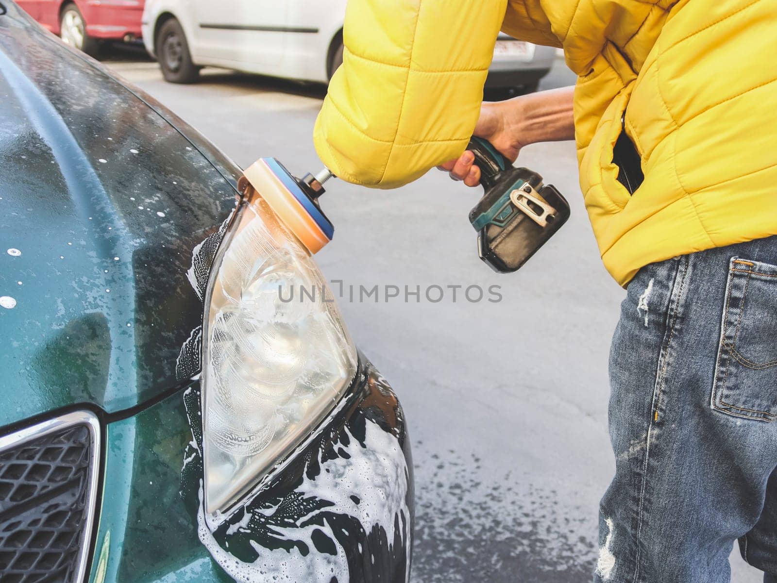A young caucasian guy without a face in a yellow jacket is being drilled with a sponge disc and polishing the headlight by Nataliya