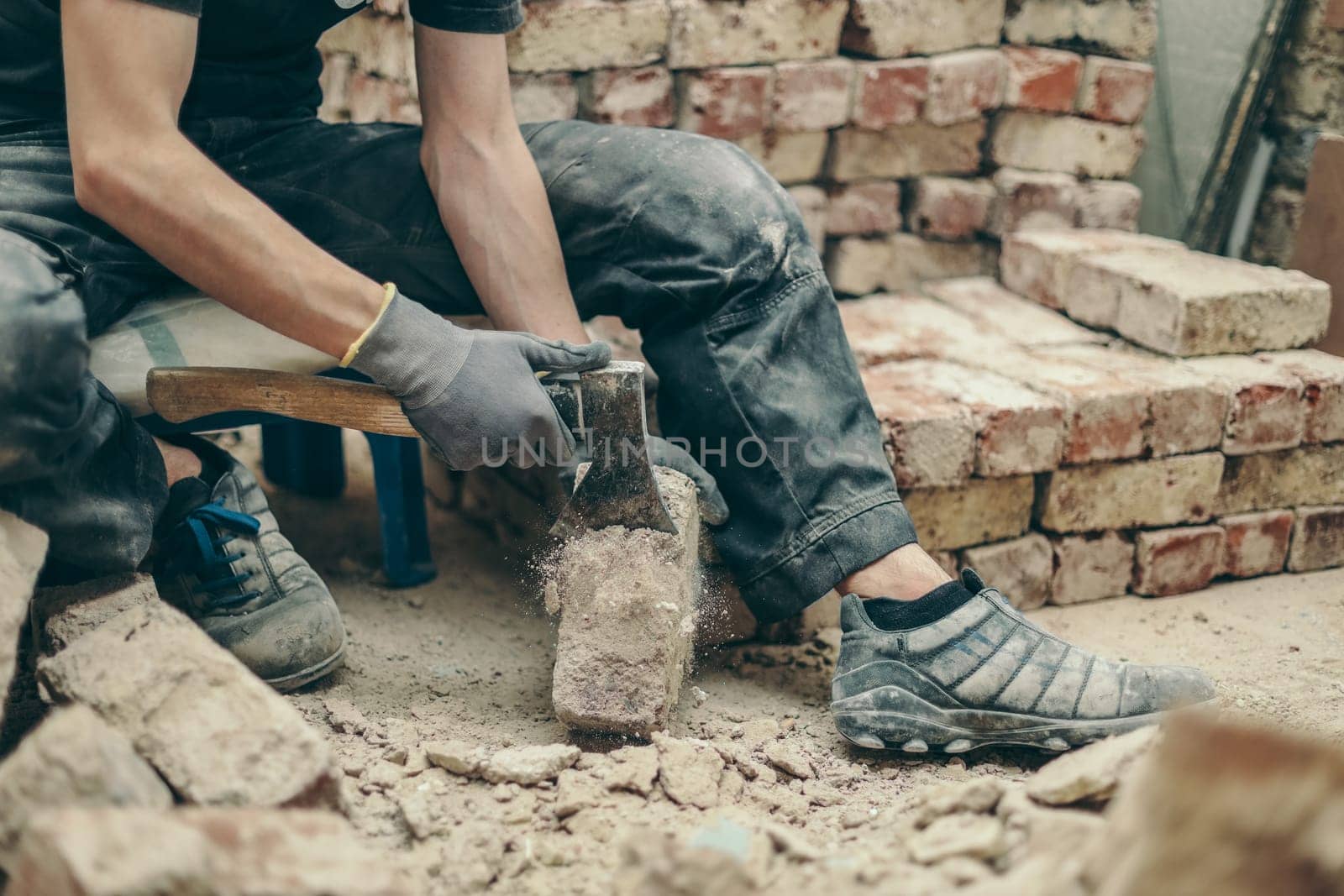 A young Caucasian unrecognizable guy in a black uniform is cleaning a dirty brick of cement residue with an axe corner, sitting on a small bench with his legs spread in a room where a pile of bricks is being repaired. Side view