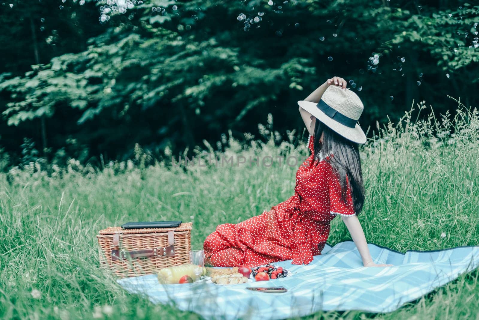 A beautiful young caucasian girl in a red dress covering her face with a straw hat sits half-turned on a blue bedspread with a wicker basket and laid out food in a clearing in a public park, close-up side view. Outdoor picnic concept.