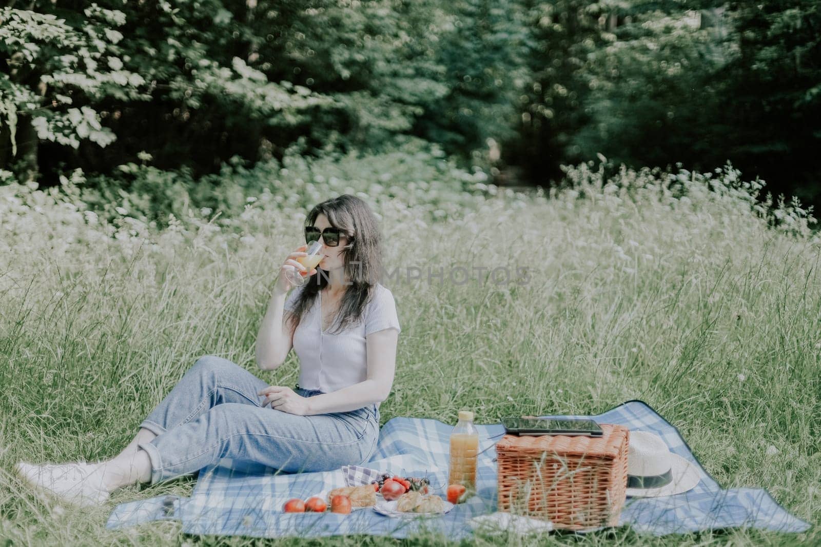 A young beautiful caucasian girl in sunglasses with brown hair, jeans and a t-shirt sits on the floor with a wicker basket, fruits in a plate and seductively drinks fresh orange juice from a glass against the backdrop of tall grasses and trees in a public park, close-up view. parks and forests, actually eat, a healthy lifestyle, lonely rest, healthy food and drinks.