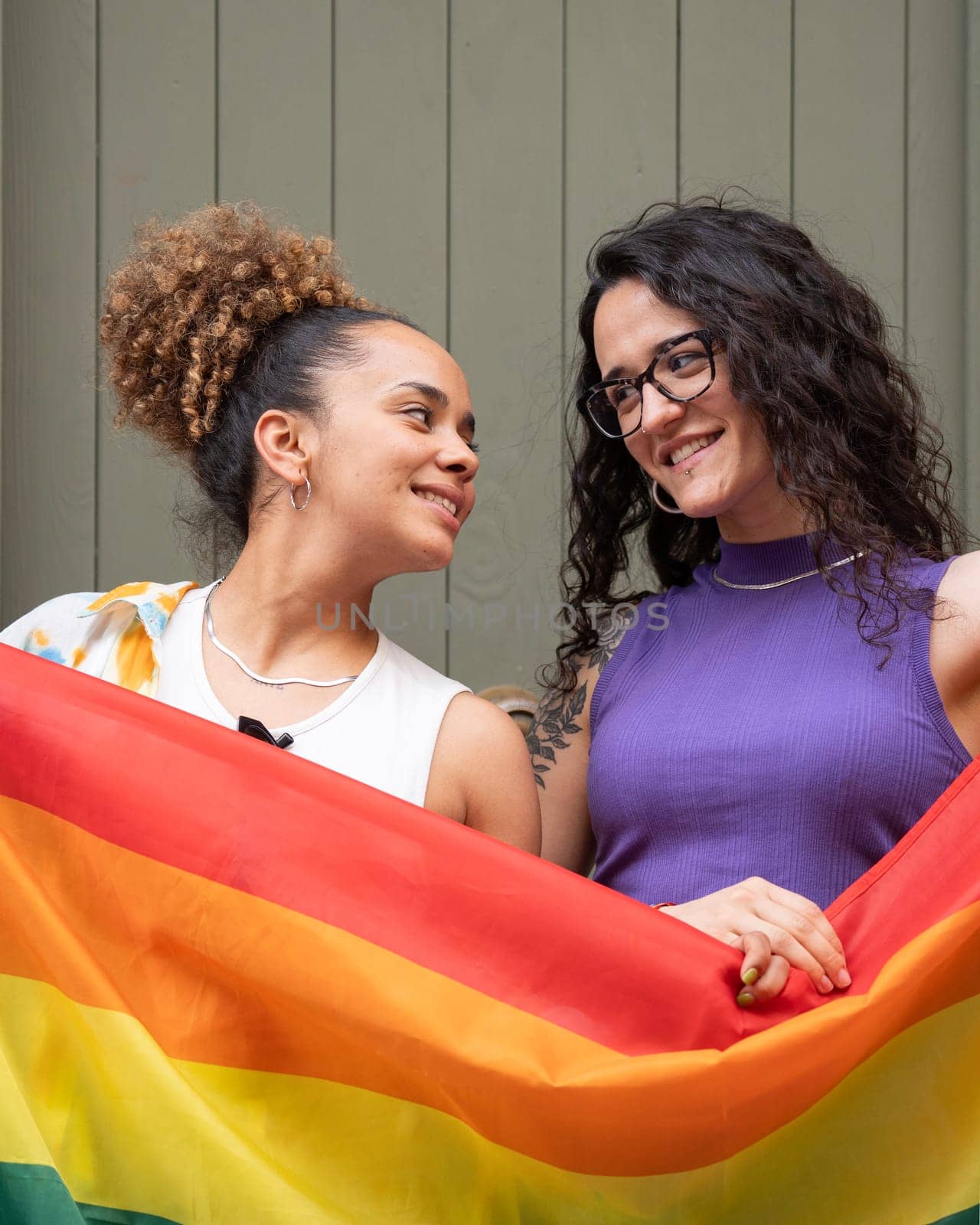 Two lesbian smiling women are holding a rainbow flag representing the LGBTQ community by papatonic