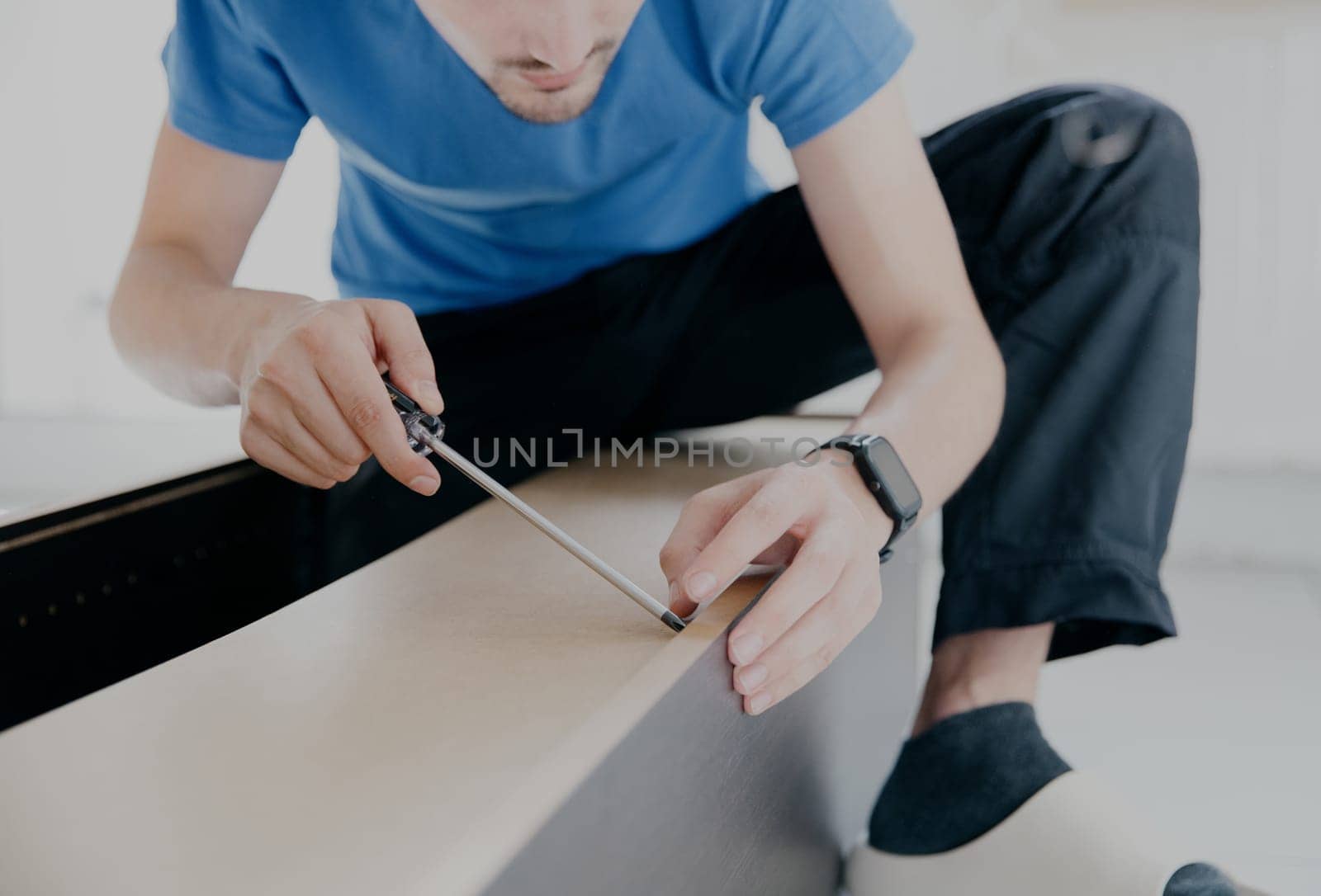 One young Caucasian man in a blue T-shirt and black pants screws a screw into the broken back wall of an old wardrobe with a screwdriver, close-up side view.