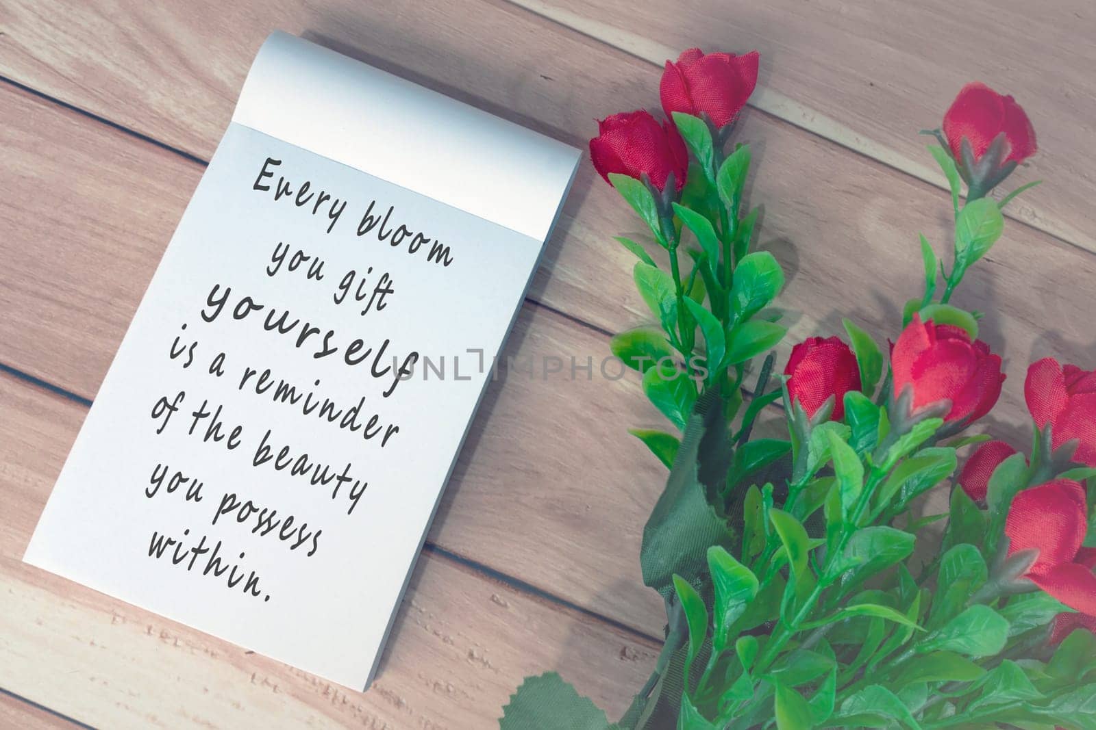 Motivational quote written on note book with artificial flowers on wooden desk by JennMiranda