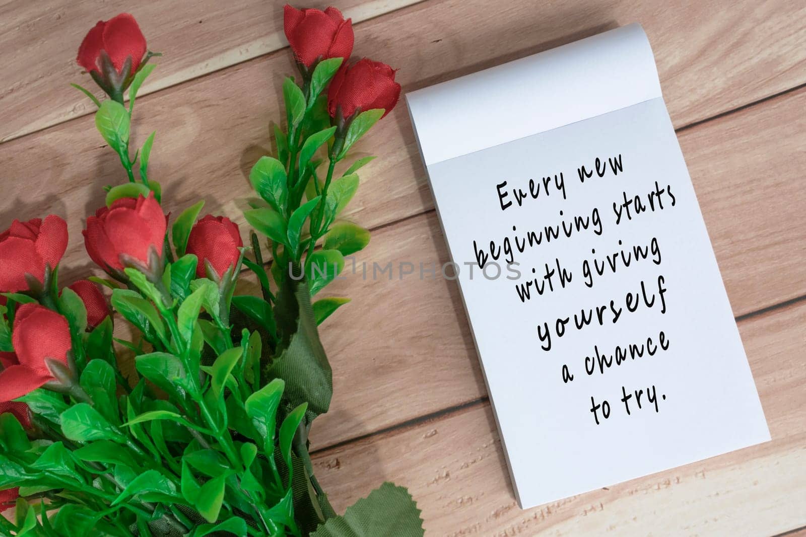 Motivational quote written on note book with artificial flowers on wooden desk by JennMiranda