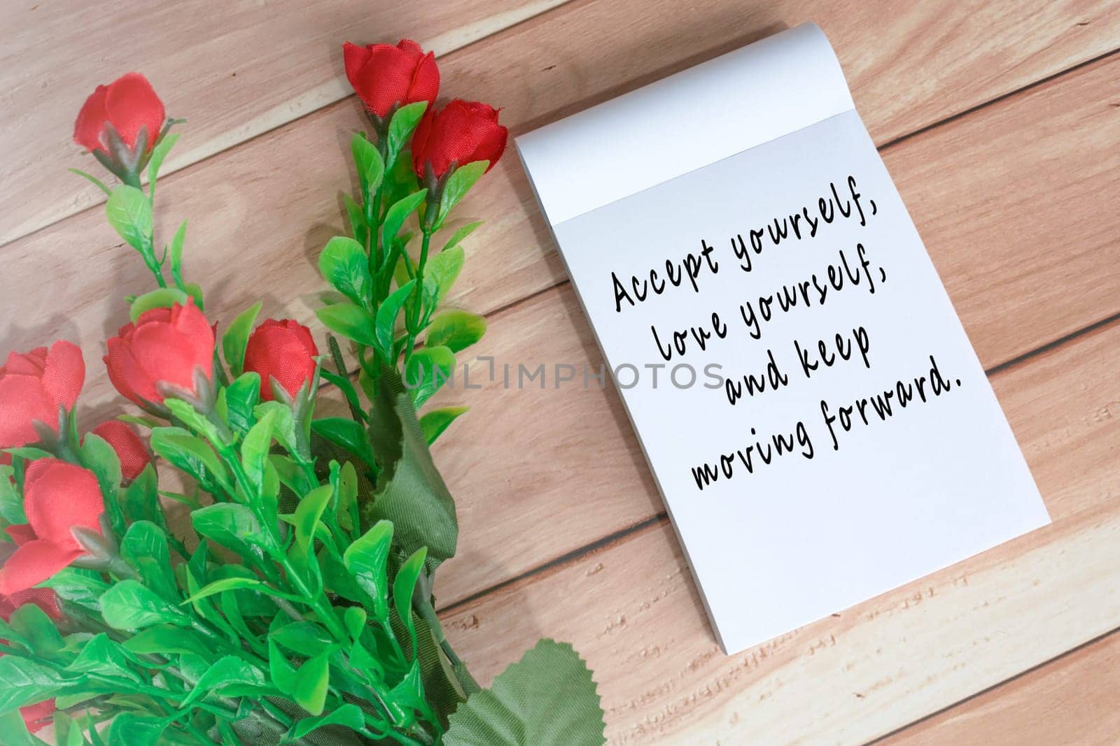 Motivational quote written on note book with artificial flowers on wooden desk by JennMiranda
