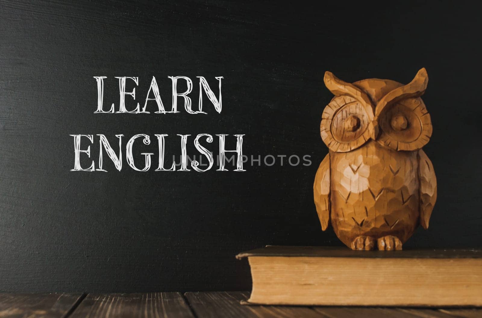 A wooden owl sits on a book next to a chalkboard that says Learn English. The owl is a symbol of wisdom and knowledge, and the chalkboard suggests that the owl is there to help people learn English