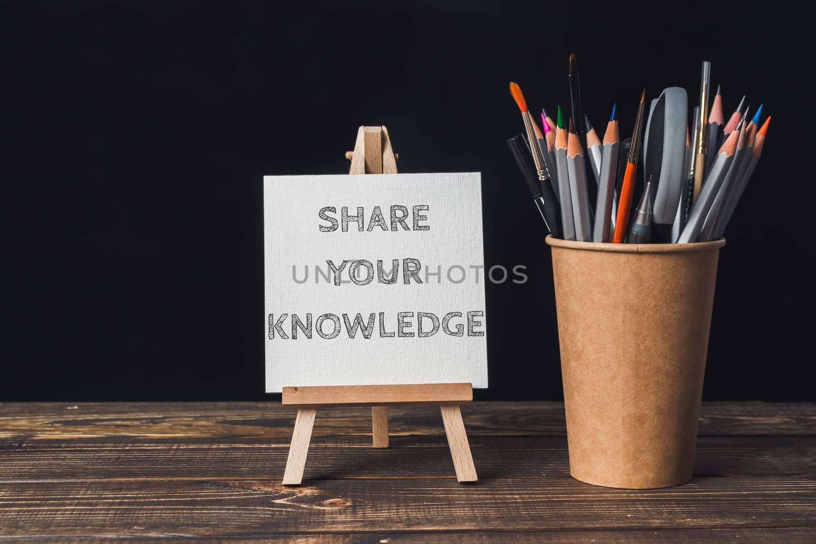 A white sign that says share your knowledge is on a wooden stand. A cup of pencils is next to the sign