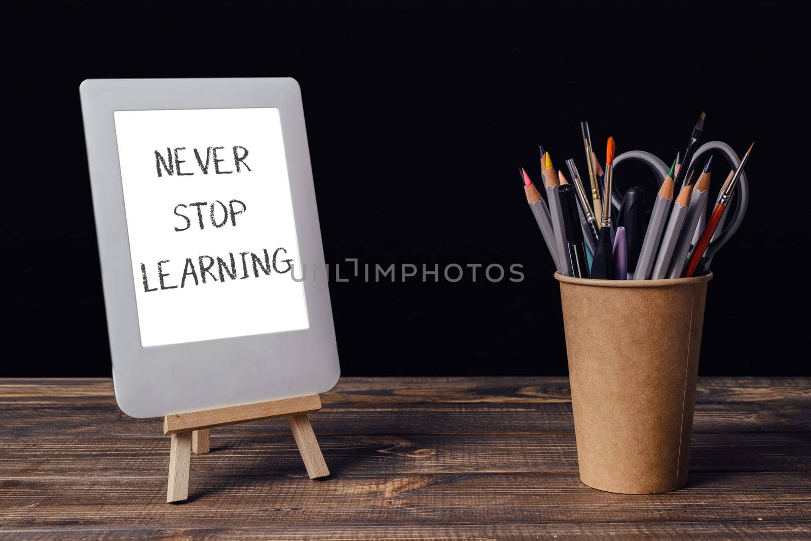 Never stop learning is written on a white sign. A wooden stand holds a white tablet and a cup of pencils
