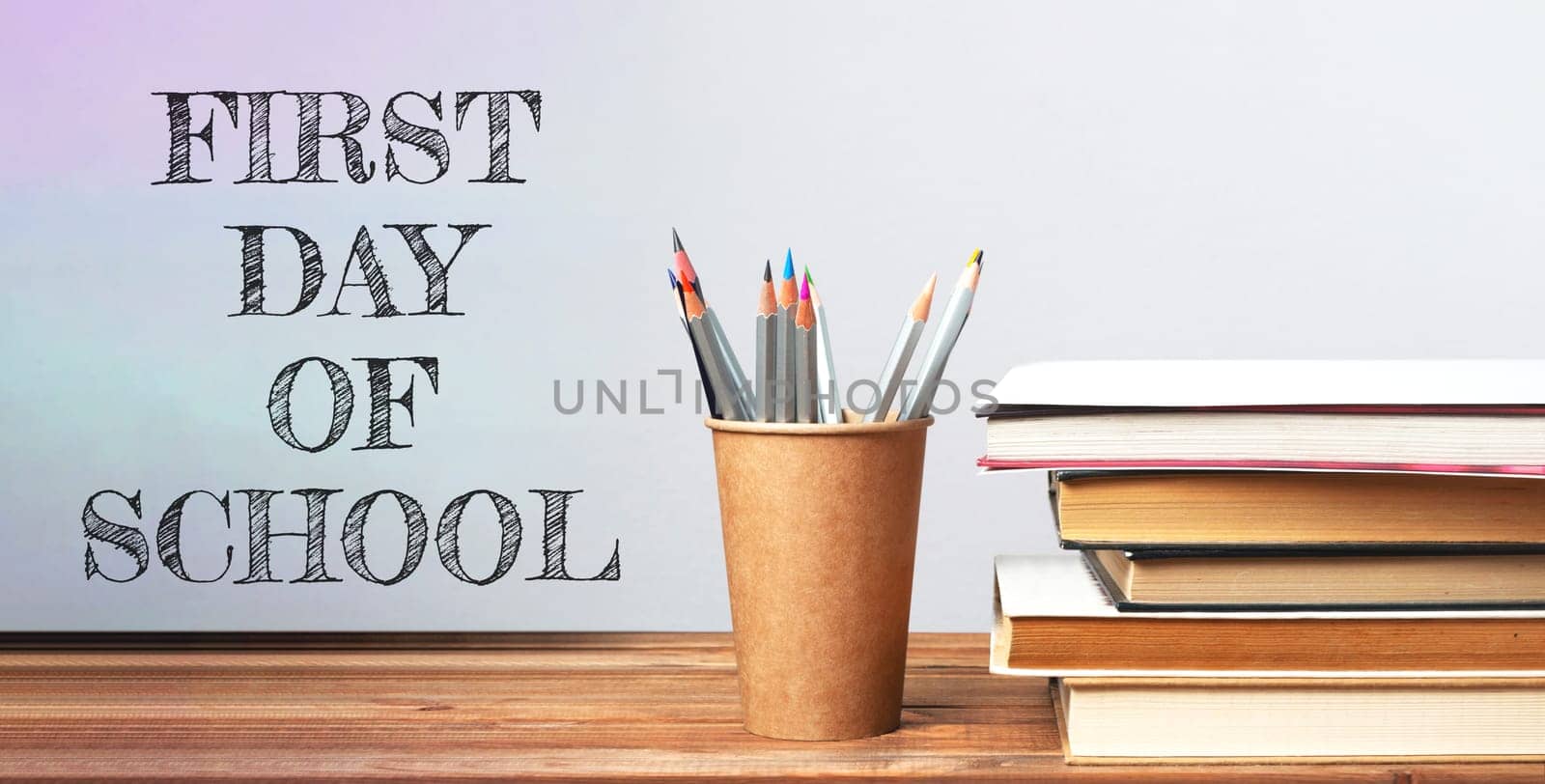 A wooden desk with a cup of pencils and a stack of books. The words First Day of School are written on the desk