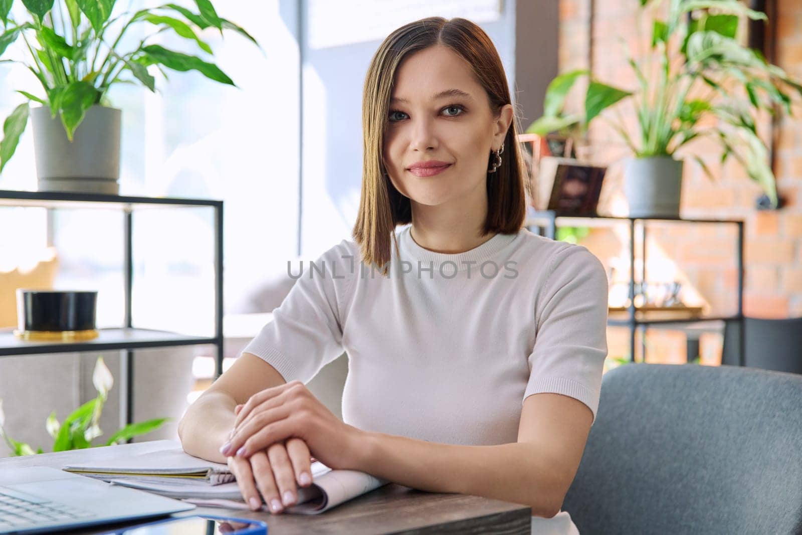 Portrait of beautiful smiling young woman sitting at table in coworking cafe by VH-studio