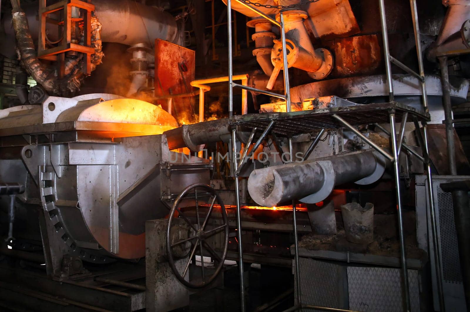 Molten metal is being poured from a ladle into a mold in a steel mill. The factory is filled with heavy industry machinery and equipment. This is a metallurgy and metalworking theme.