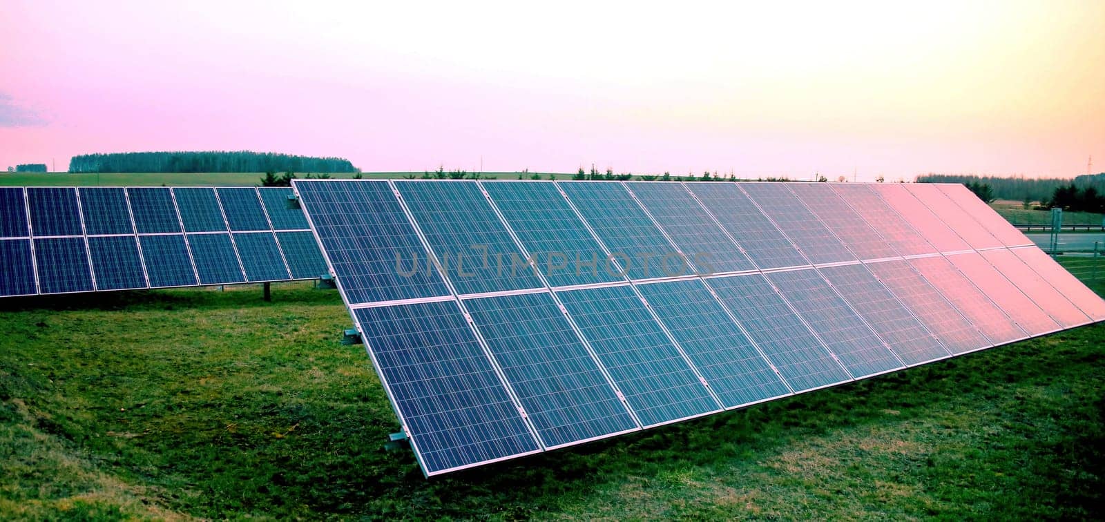 Rows of blue photovoltaic panels stand in an open field, capturing the last light as the sun sets on the horizon. The sky displays a gradient of colors from blue to soft orange, signifying the transition from day to night. The solar farm generates sustainable energy in a rural setting, highlighting the blend of technology with the natural environment.