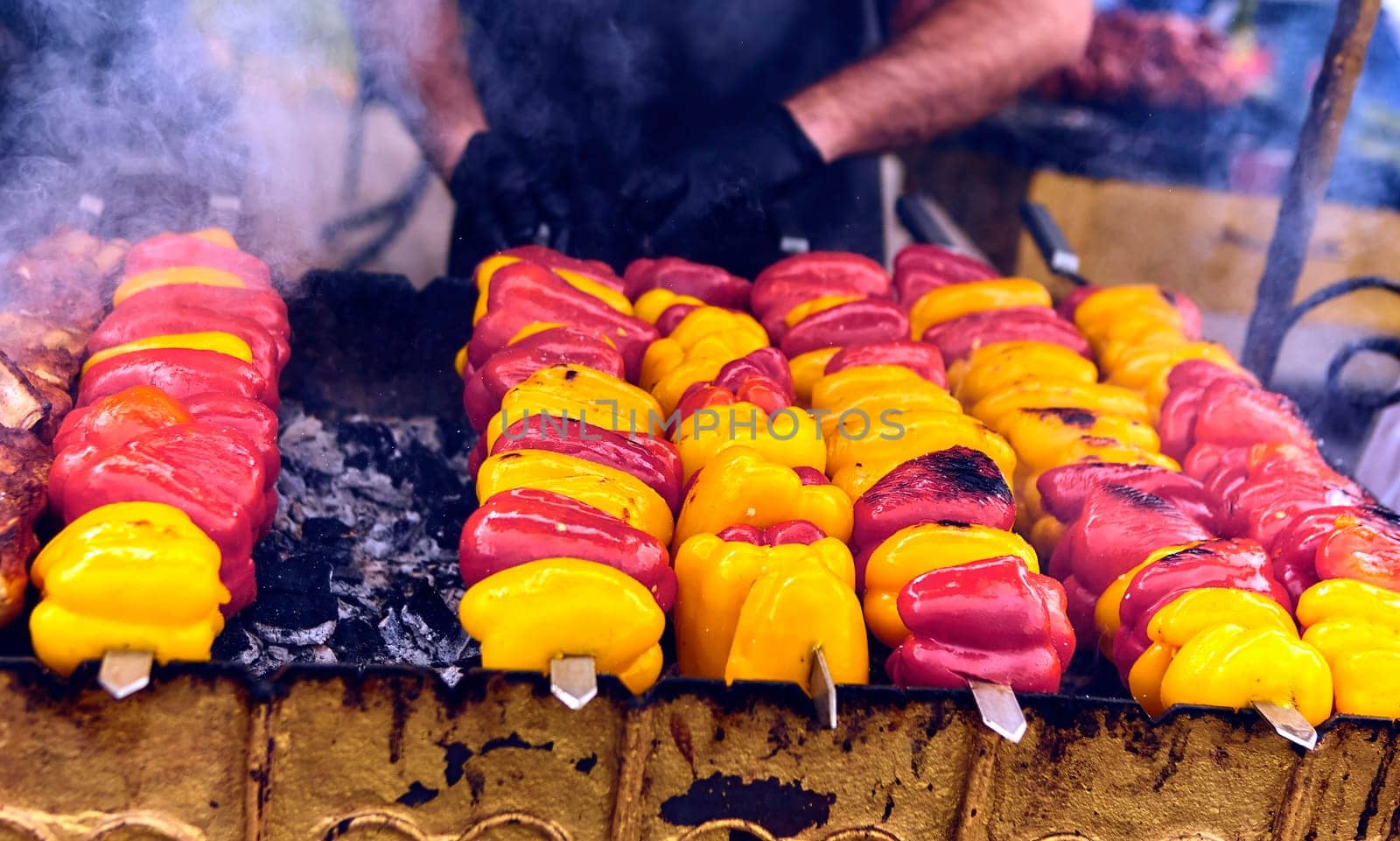 Rows of colorful bell peppers on skewers being grilled on a large outdoor grill by Hil