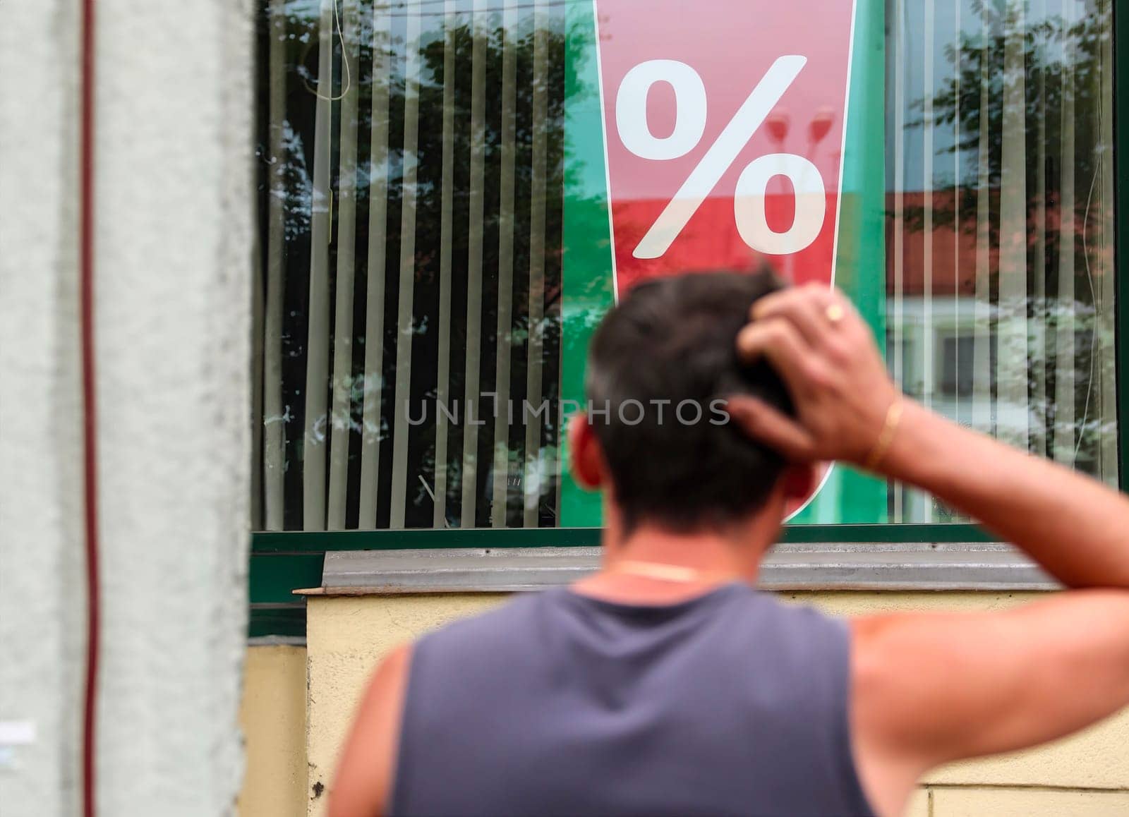 A man is standing outside a store, scratching his head as he observes a large percentage sign displayed in the window. His posture suggests he might be puzzled or considering the implications of the advertised discount, sale, or promotion.