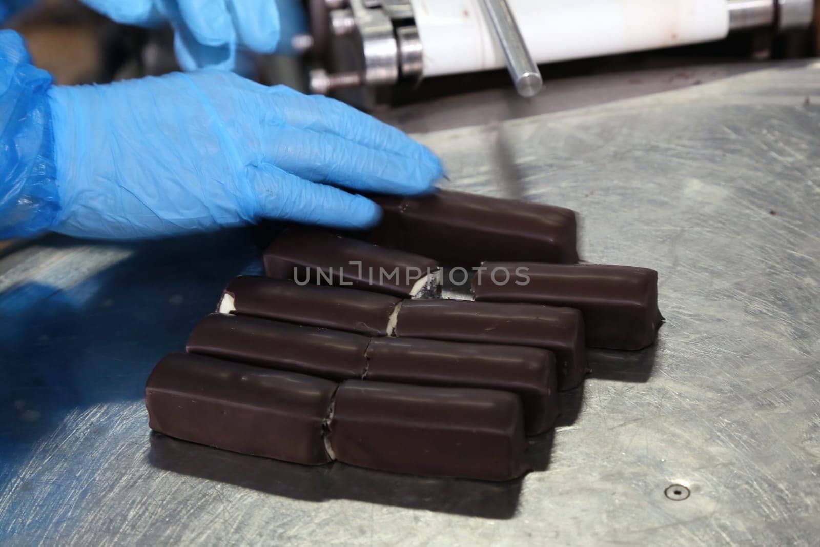 A gloved hand inspects a batch of freshly made chocolate bars on a stainless steel surface at a confectionery production facility. Each bar is aligned uniformly, awaiting the next phase of packaging or additional quality checks to ensure consistency and excellence.