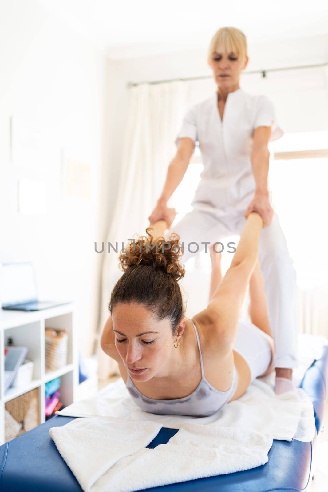Professional osteopath pulling hands of woman lying on stomach while fixing shoulder joint during rehabilitation session