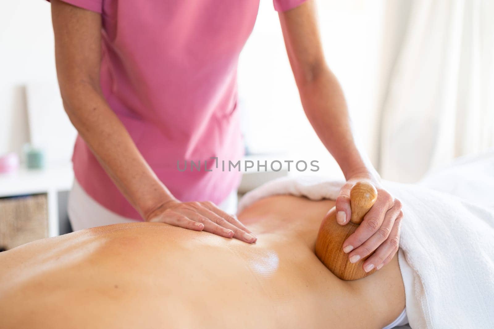 Crop masseur massaging back of anonymous customer with wooden massage therapist tool during physiotherapy treatment in modern clinic