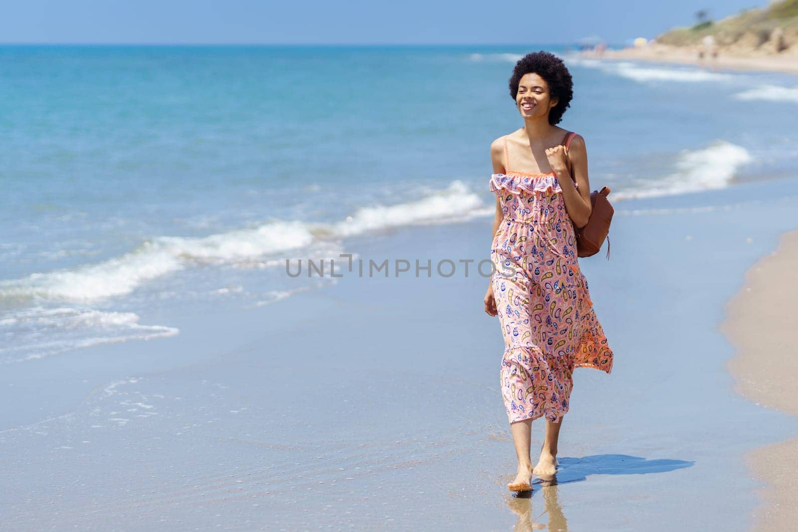 Full body of young African American female in colorful dress smiling while spending summer vacation on sandy beach near rippling sea and looking away