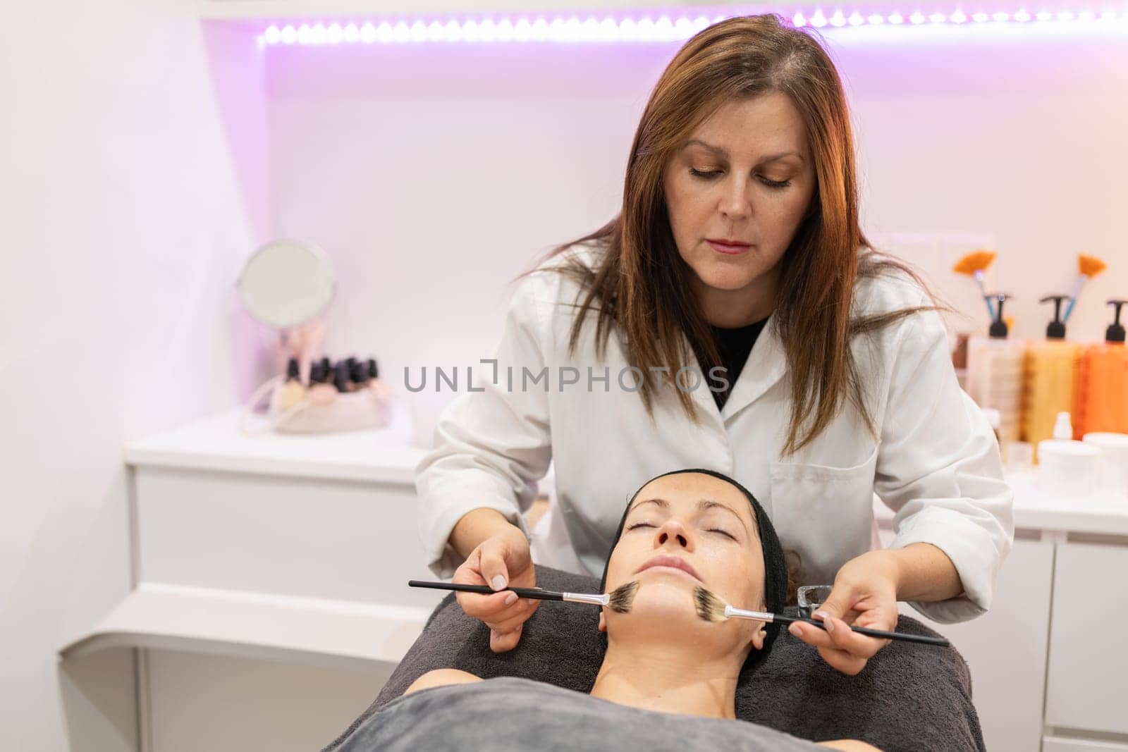 Beautician applying facial cleansing oil, with brush on woman lying face up with eyes closed during cosmetology session in beauty salon in background with cosmetics on counter