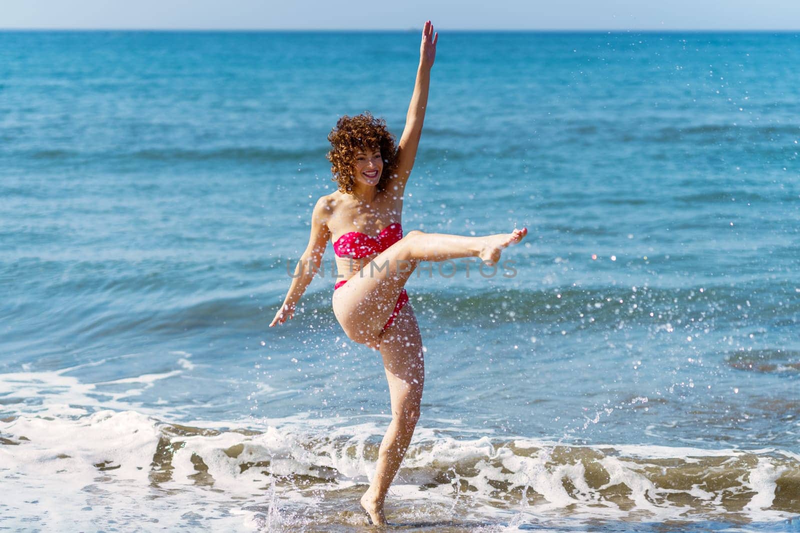 Full body of playful female in bikini kicking sea water and splashing on beach while enjoying sunny summer day