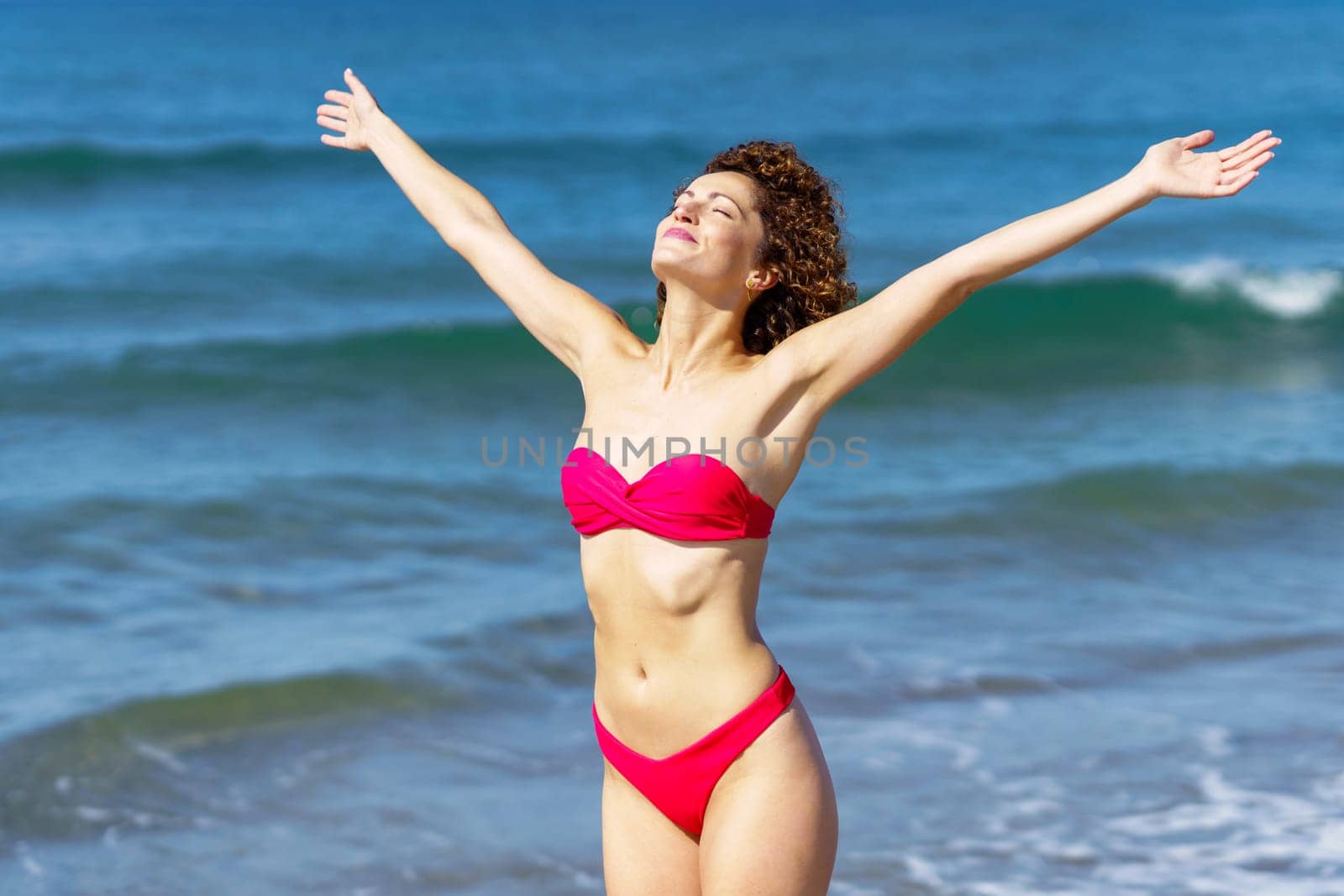 Delighted female with closed eyes in pink swimwear raising arms while sunbathing and relaxing against endless waving blue sea in summer