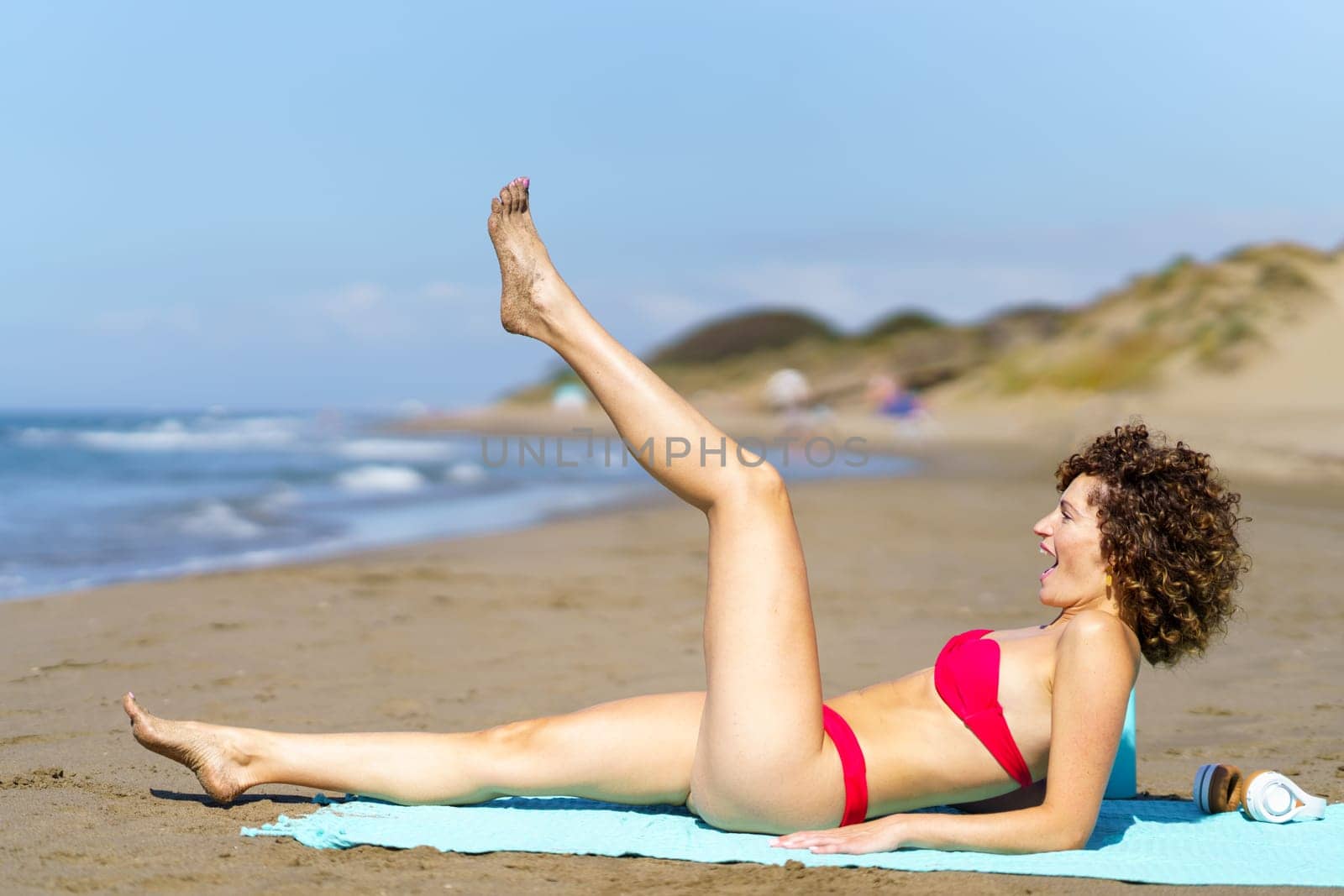 Side view of young woman with curly hair in pink bikini raising leg while having fun on sandy shoreline