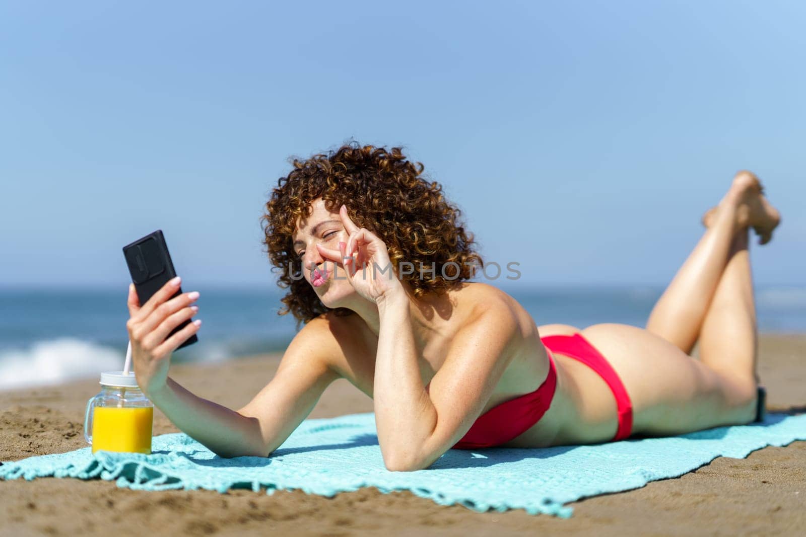 Young curly haired female in bikini lying on shore of ocean and showing two fingers while taking selfie in sunlight