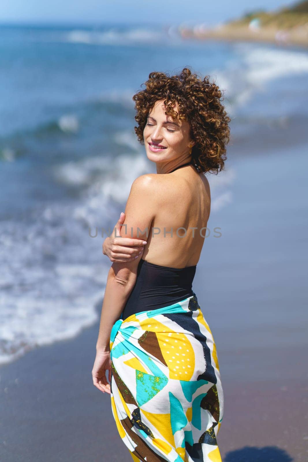 Side view of young sensual female in casual colorful clothes with curly hair standing on seashore with closed eyes while enjoying outdoor breeze in daylight