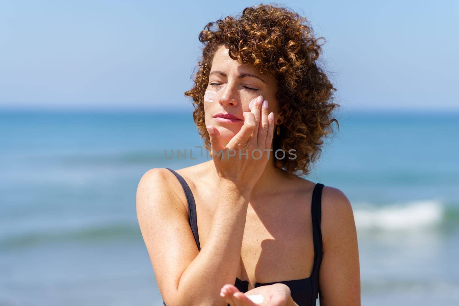 Young curly haired female in black bikini applying sunscreen cream on cheeks while standing near sea on sunny summer day protecting skin from sunburn