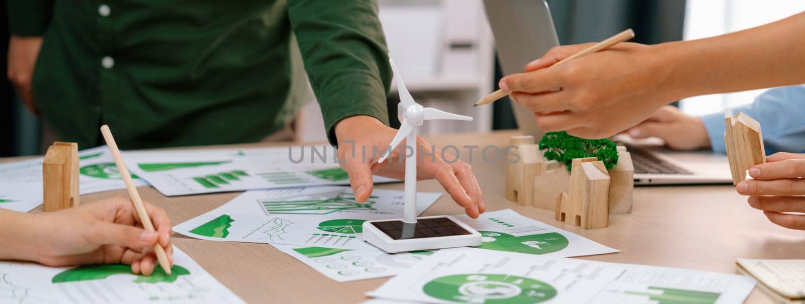 Windmill model represented renewable energy and wooden block represented eco city was placed on green business meeting table with environmental document scatter around. Front view. Delineation.