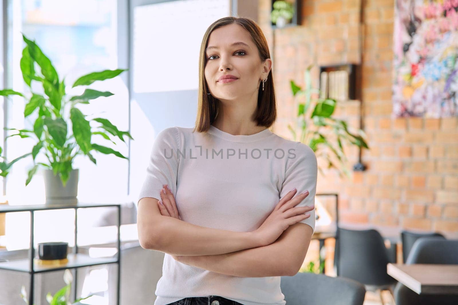 Portrait of young confident beautiful woman with crossed arms looking at camera in coworking cafe. 20s female university college student, businesswoman, freelancer employee, young specialist