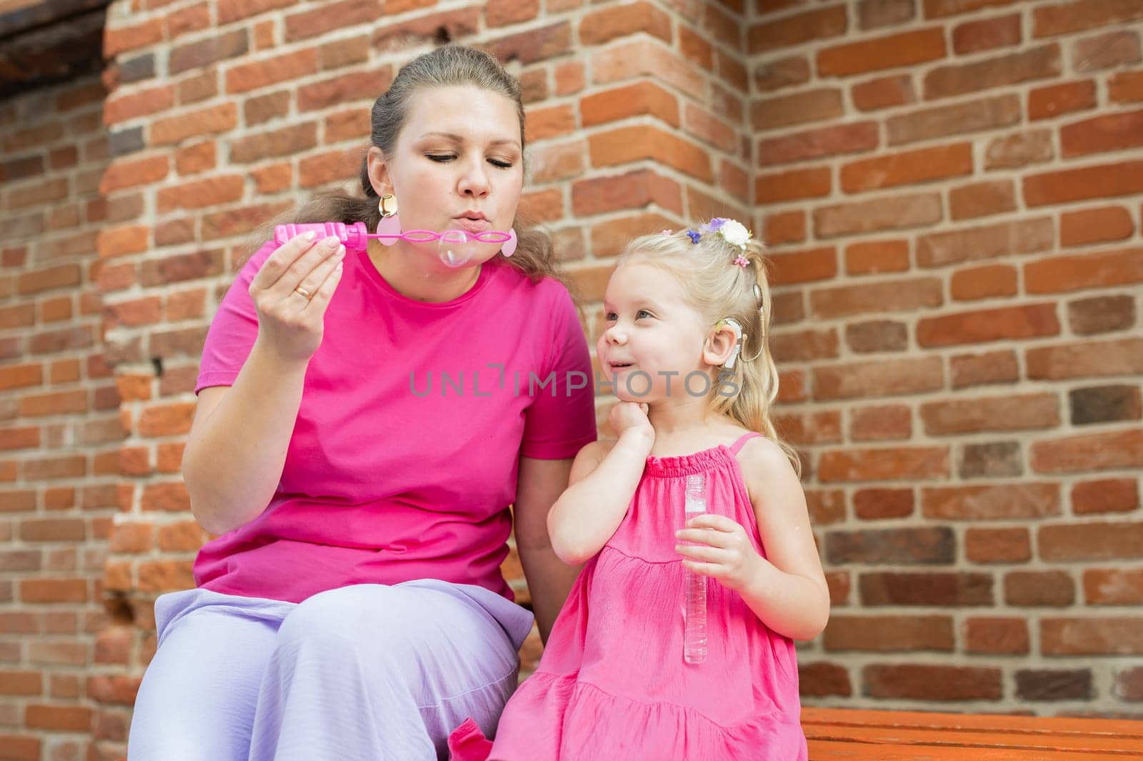 Blonde little girl with cochlear implant blowing soap bubbles with her mother outdoor. Hear impairment deaf and health concept. Diversity and inclusion. Copy space by Satura86