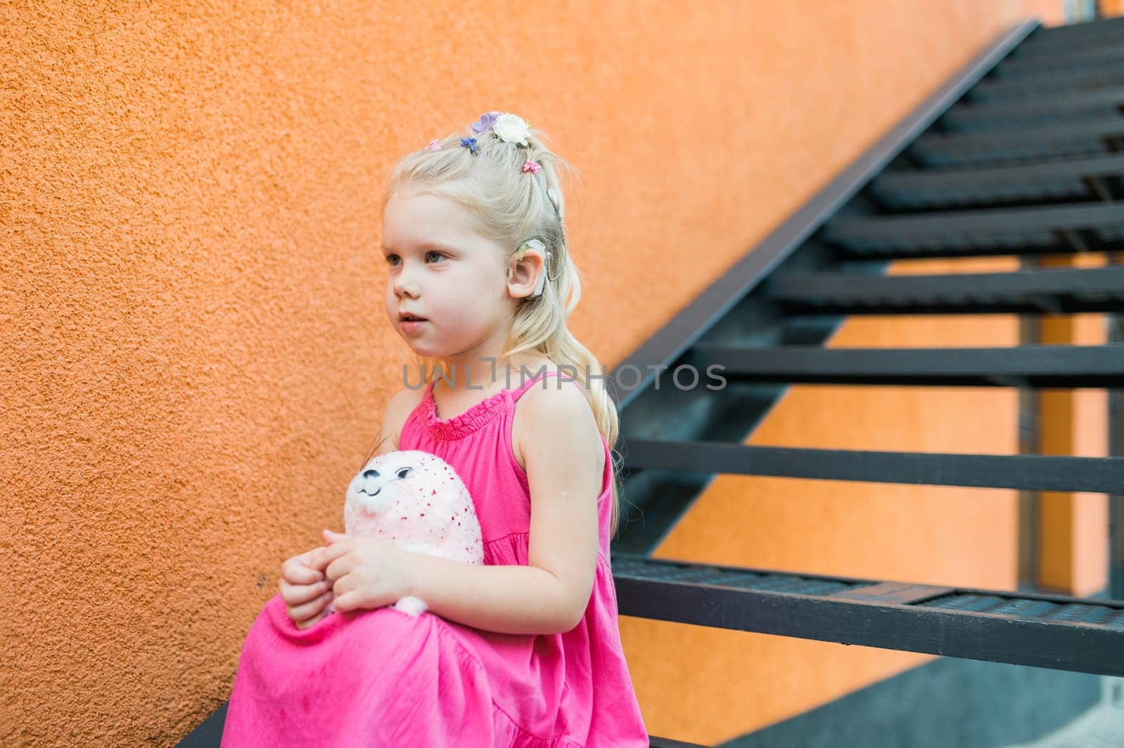Child girl with hearing aid cochlear implant in pink dress having fun on summer street. Aid for the treatment of deafness and hearing loss in humans. Copy space by Satura86