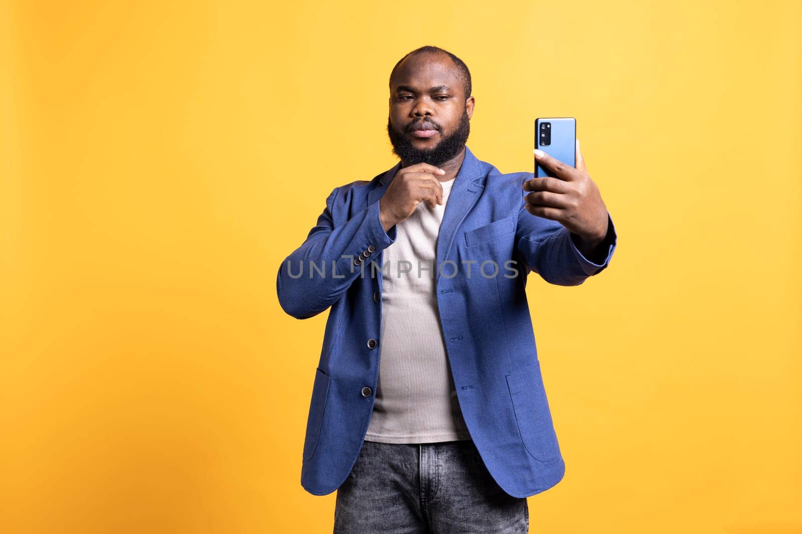 Narcissistic man using cellphone to take selfies, stroking his chin. Vain social media user taking photos using phone selfie camera, isolated over yellow studio background