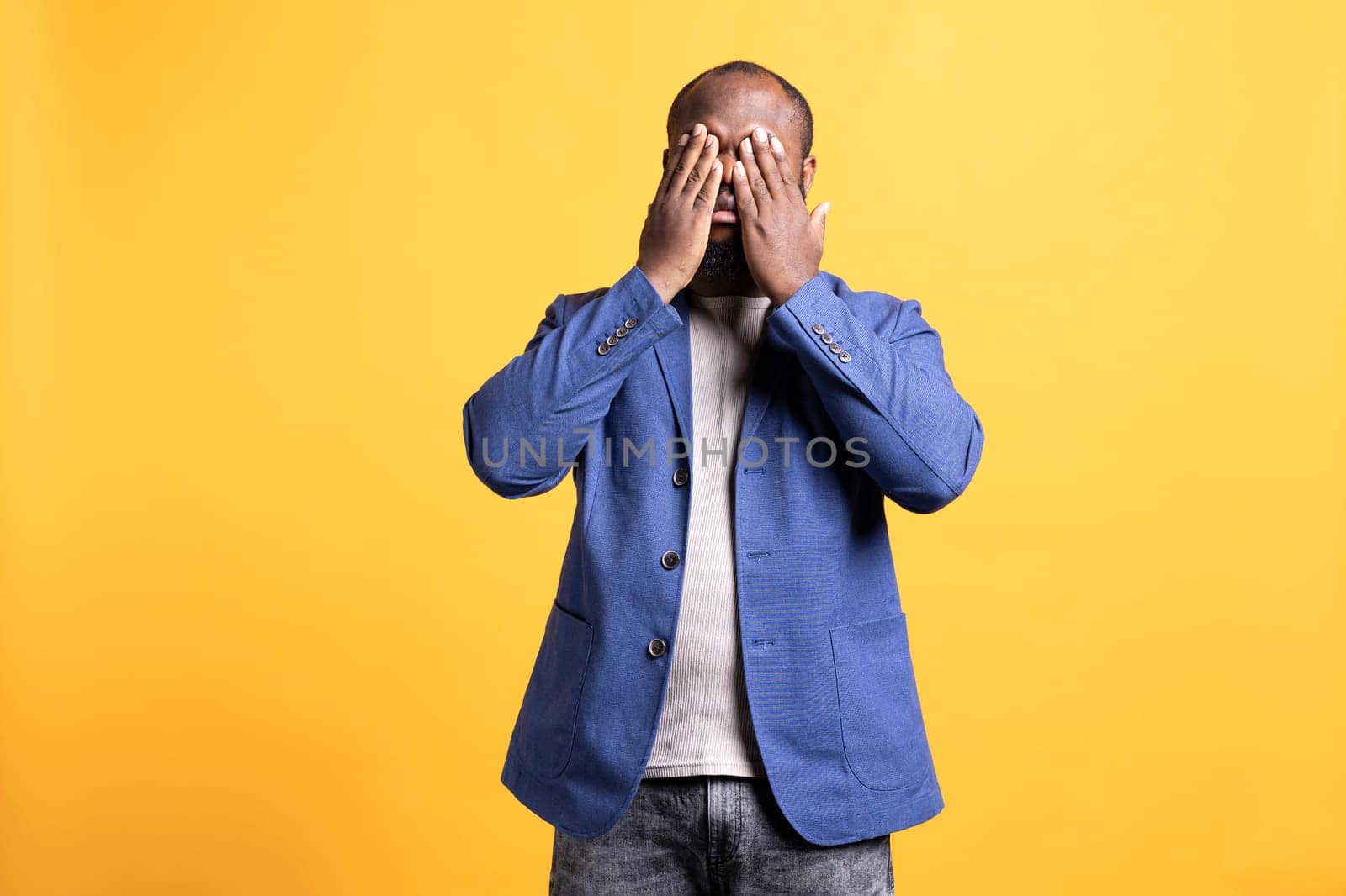 Tired BIPOC man covering face with palms, exhausted after long day at work. African american person in need of break from job, obstructing eyes with hand, isolated over studio background