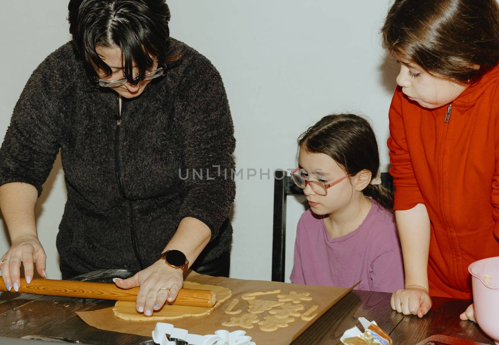 Two beautiful Caucasian brunette girls are sitting at the table and together they carefully watch how their mother rolls out dough with a rolling pin, close-up side view. Step by step instructions. Step 18