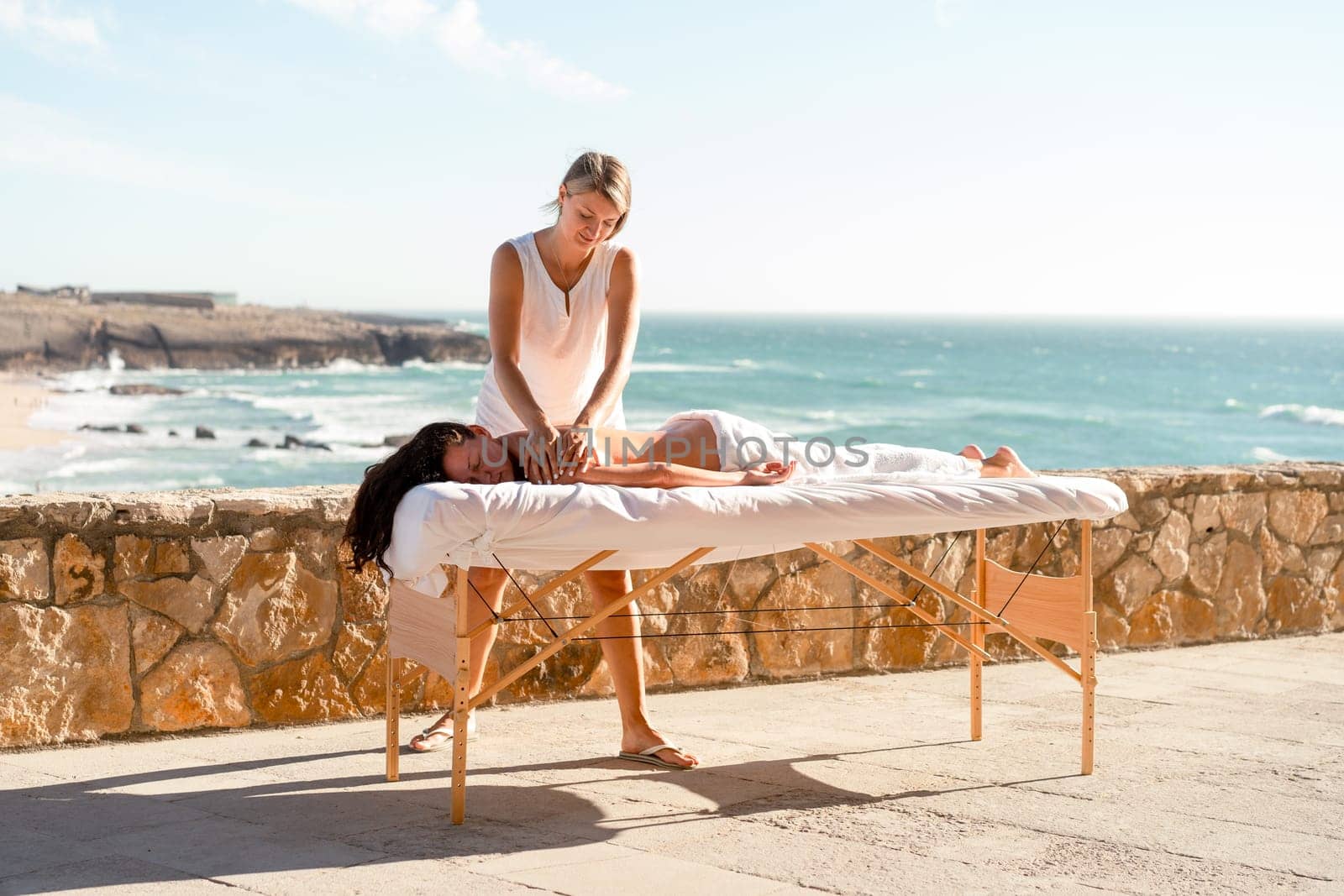 Tanned brunette woman relaxing during spa treatment on ocean shore. Woman lying on massage table and getting professional back massage, beach and turquoise waves background