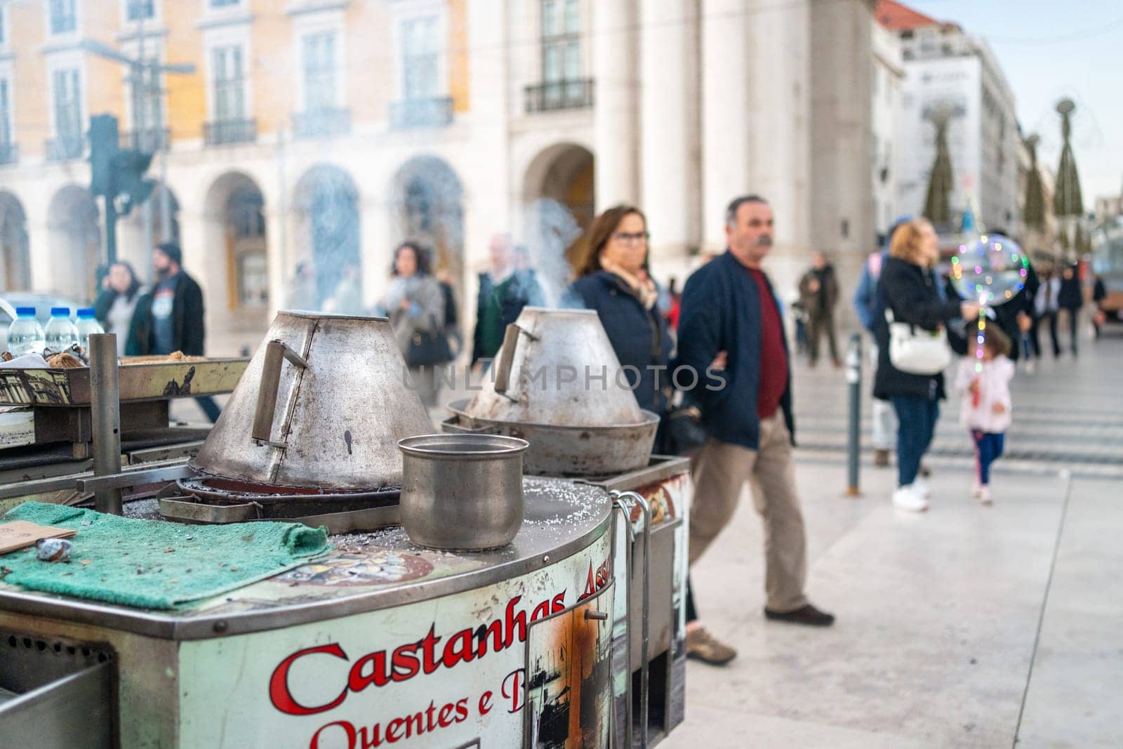 Street vendor selling roasting fresh chestnuts with smoke emitting from containers on Lisbon street. Traditional street food with blur crowd passing on Portugese city street.