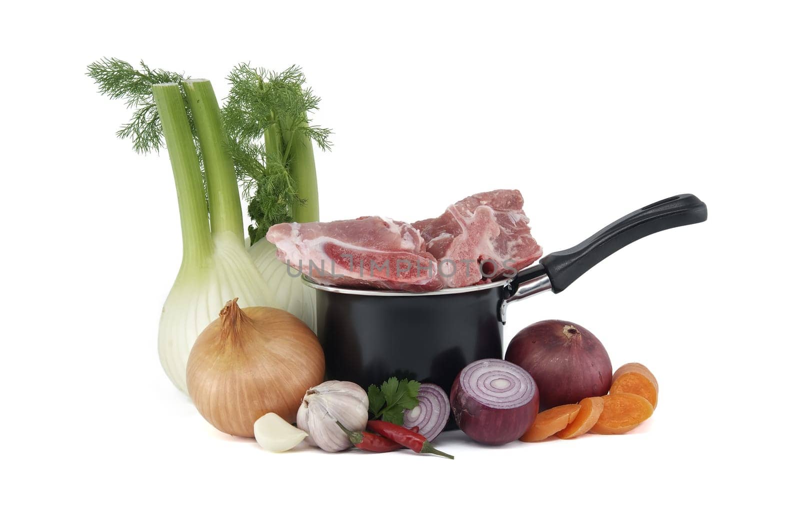 Black saucepan with pieces of raw pork atop. Around the saucepan are sliced vegetables which include onions, carrots, and garlic isolated on white background. Preparation of meat broth