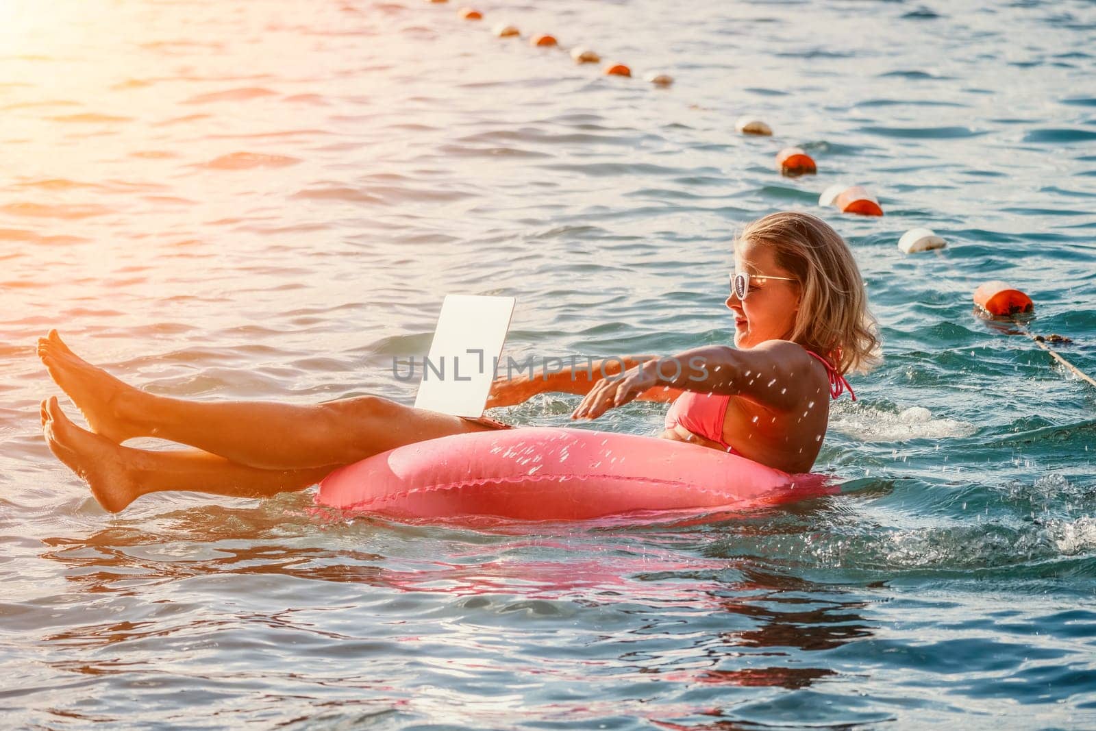 Woman works on laptop in sea. Freelancer, young blond woman in sunglases floating on an inflatable big pink donut with a laptop in the sea at sunset. Freelance, travel and holidays concept