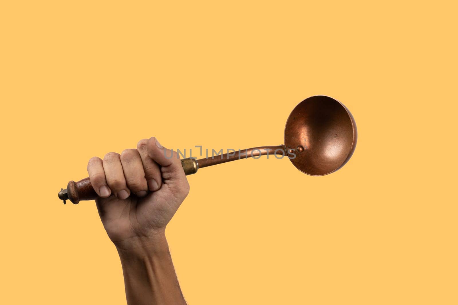 Black male hand holding a brass vintage kitchen ladle isolated yellow background by TropicalNinjaStudio