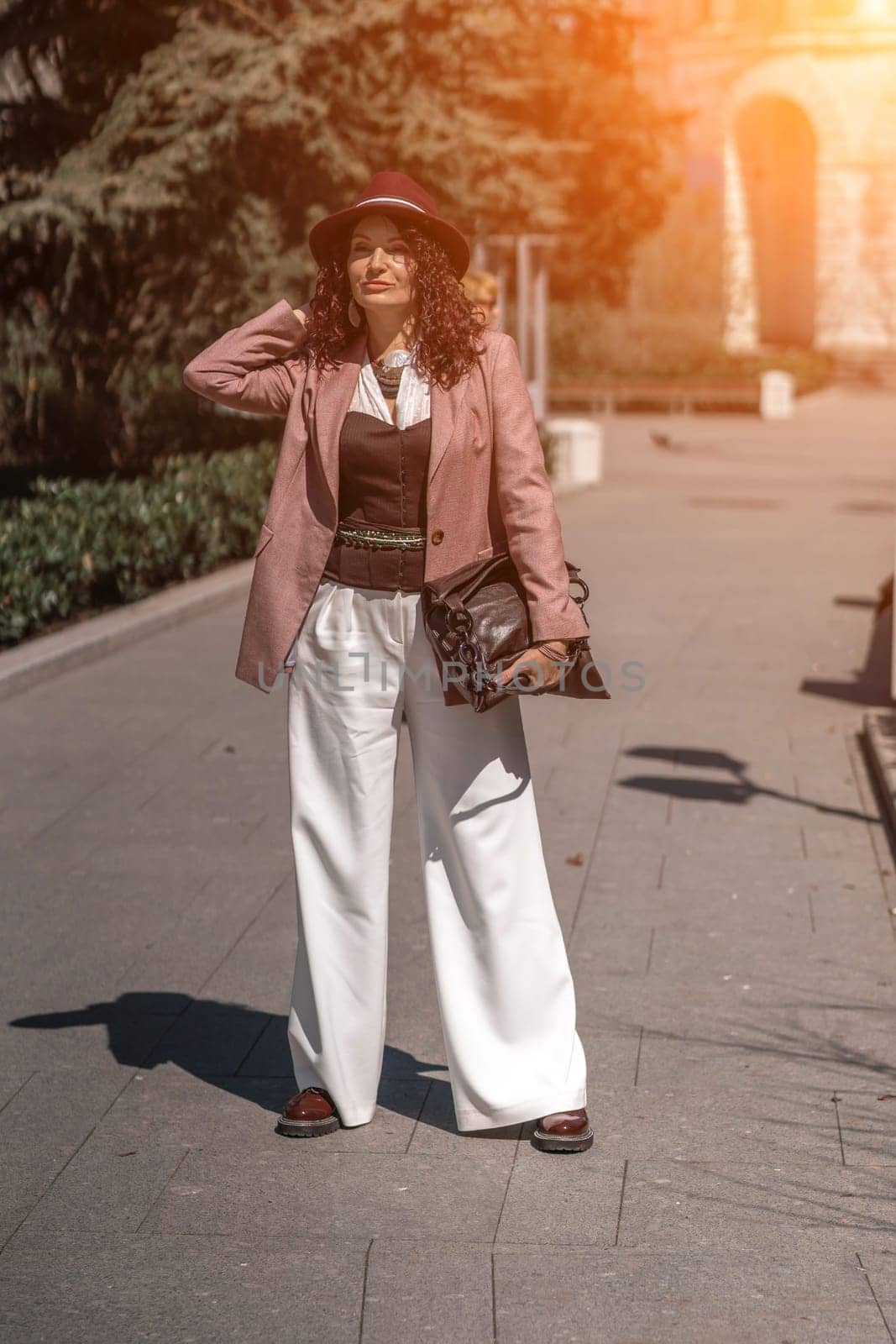 Woman park city. Stylish woman in a hat walks in a park in the city. Dressed in white corset trousers and a pink jacket with a bag in her hands