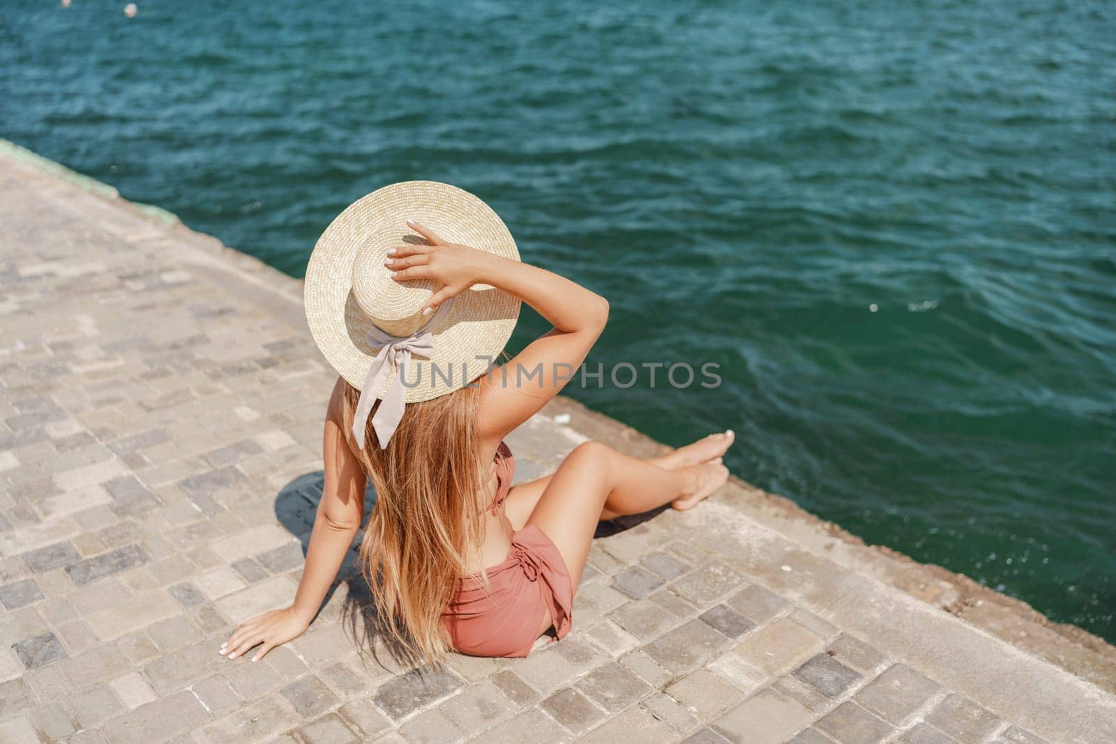 A woman in a swimsuit sits with her back holding a hat, looks at the ocean, sunny day, relaxes