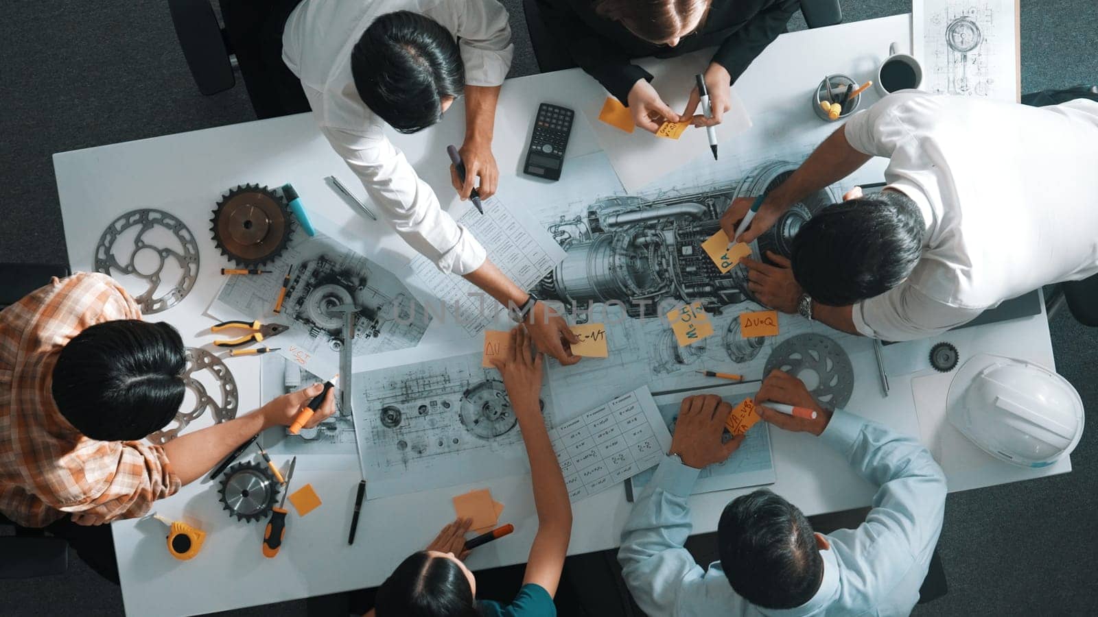 Top view of smart engineer team in casual cloth talking about turbine engine structure at table with metal gear, calculator, sticky notes. Group of technician taking a note in meeting. Alimentation.