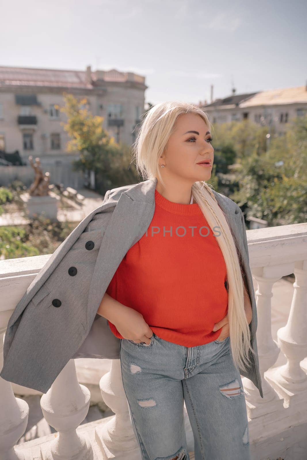 A blonde woman wearing a red shirt and gray jacket stands on a balcony. She is wearing ripped jeans and has her hands in her pockets. by Matiunina