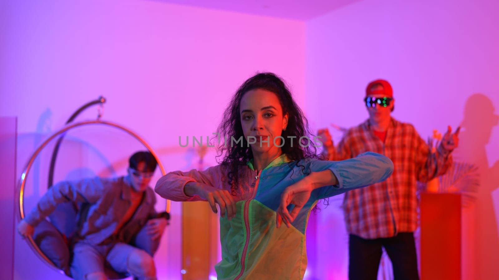 Attractive happy dancer looking at camera while smart woman moving at rhyme with neon light. Professional hispanic performer break dancing while wearing colorful cloth with diverse friend. Regalement.